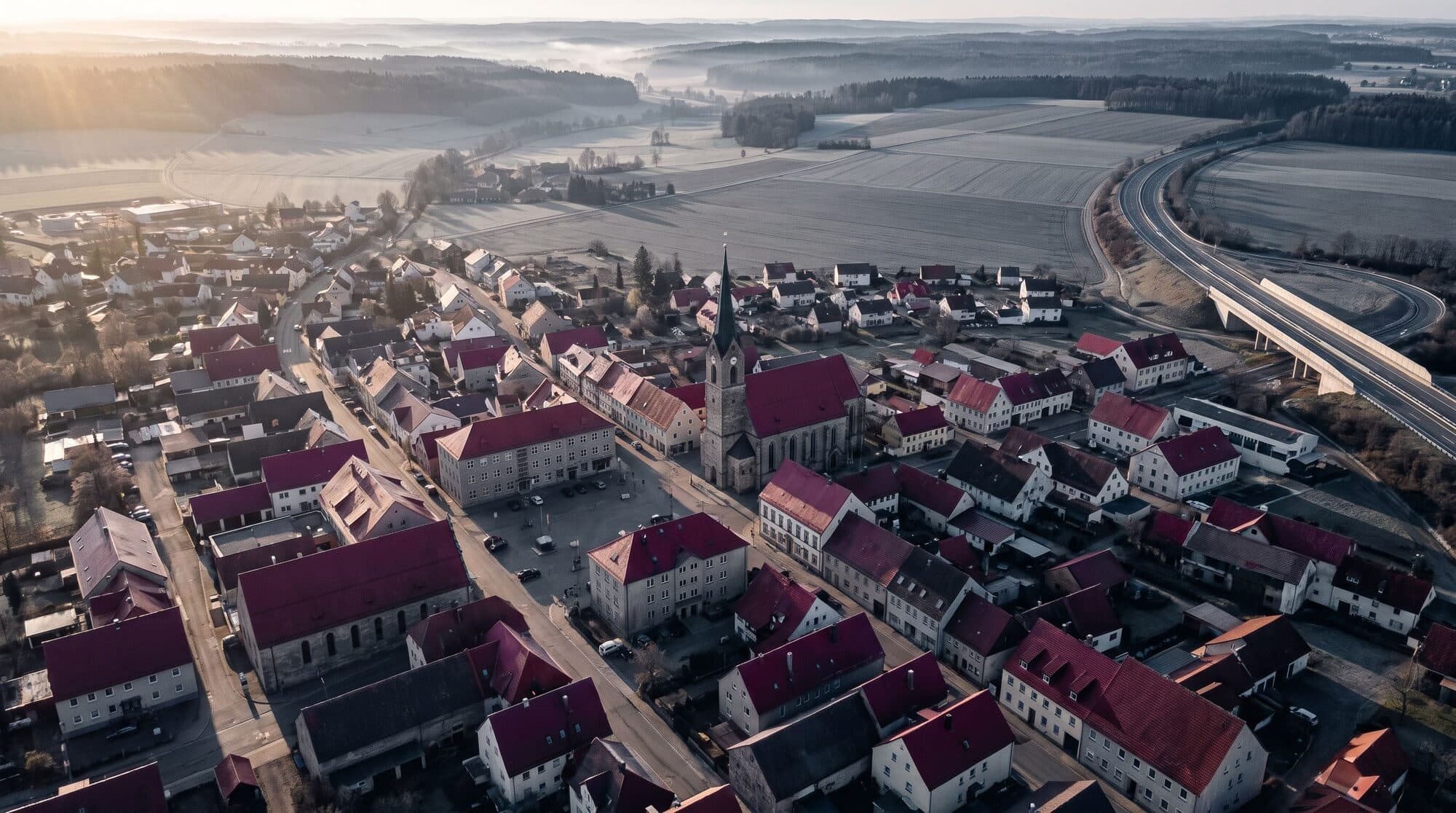Luftbild einer Kleinstadt im Morgenlicht, rote Ziegeldächer, ein Kirchturm, dahinter Felder und Waldrücken